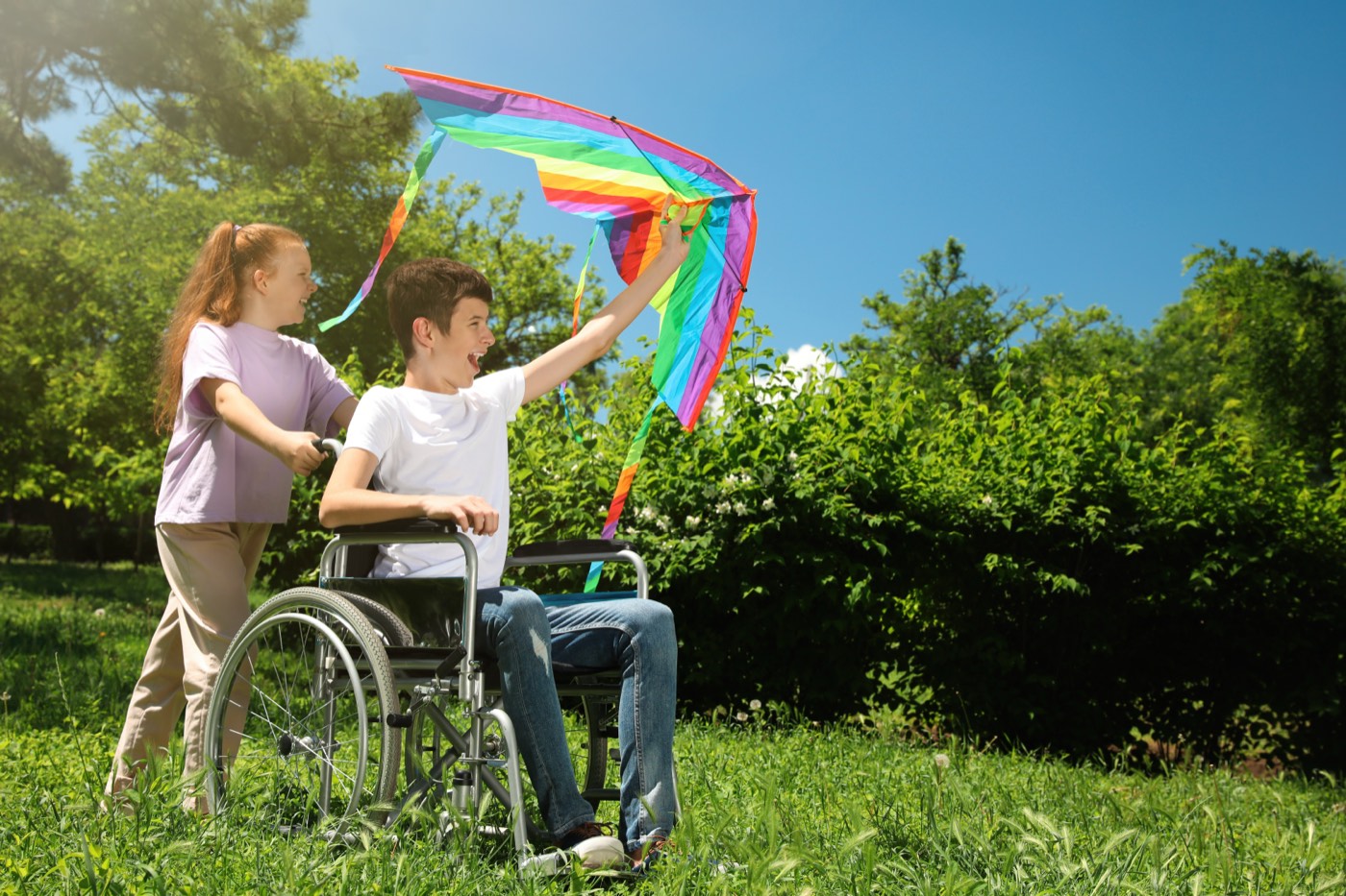 A teenage boy in a wheelchair flying a rainbow kite with a friend in a sunlit park